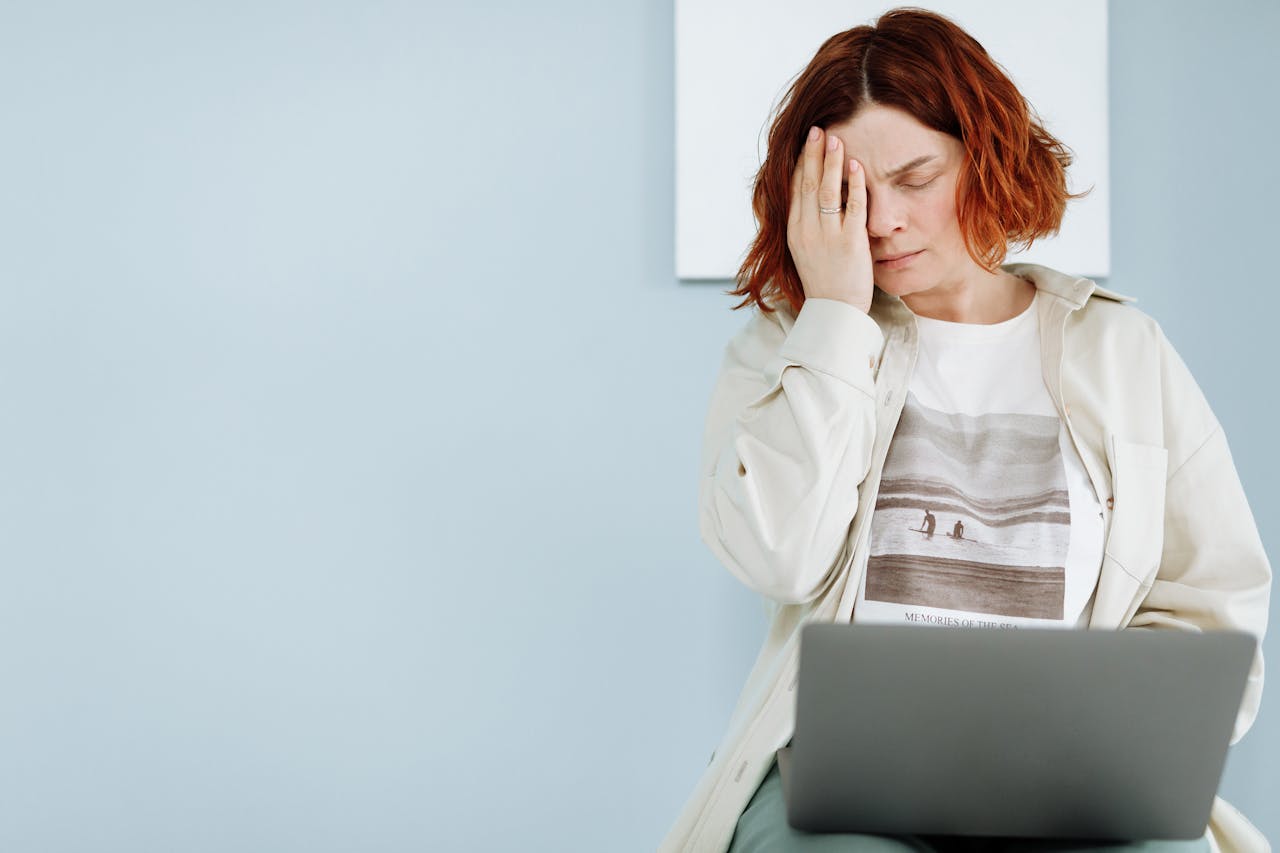 A woman experiencing stress, seated with a laptop, holding her forehead.